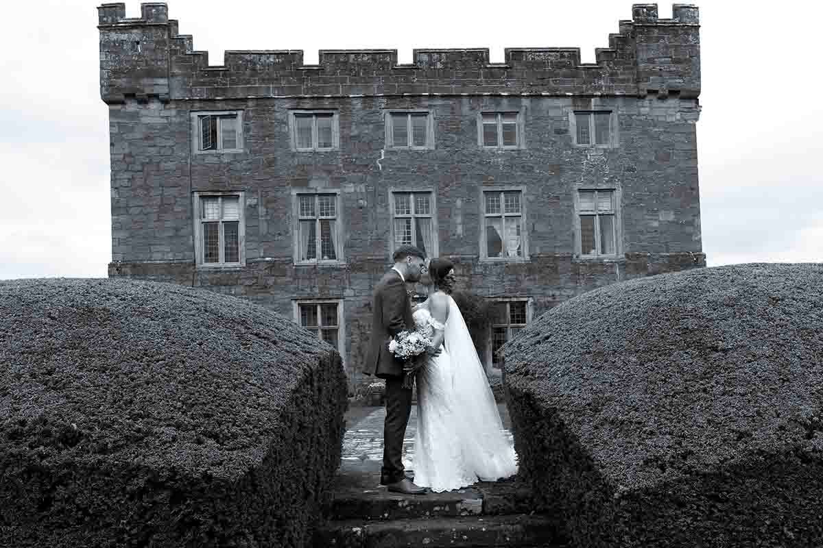 Black and white wedding photo of bride and groom kissing in front of a historic castle.