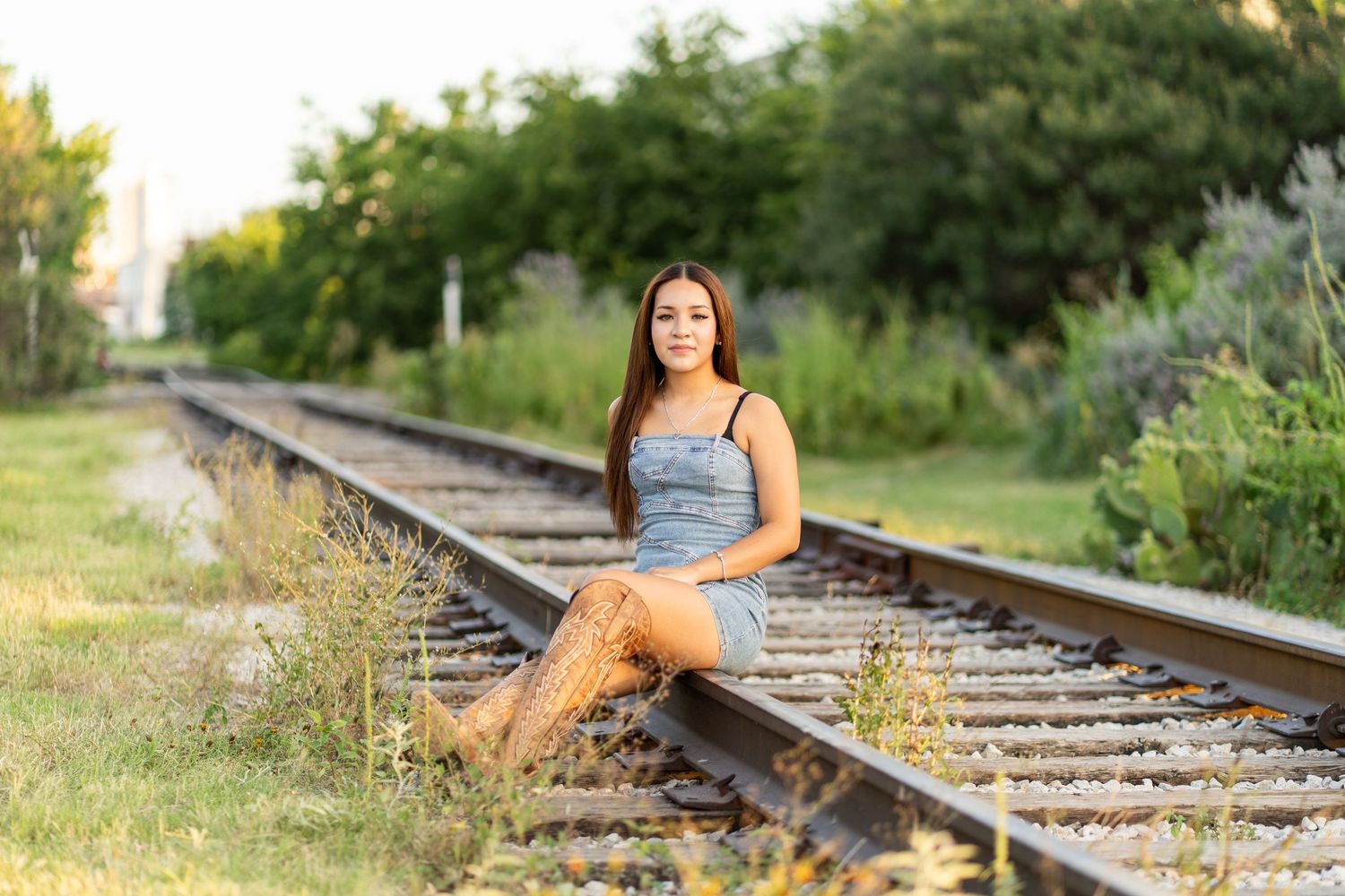 Young woman in denim dress and boots sitting on railroad tracks surrounded by lush greenery in golden hour light.