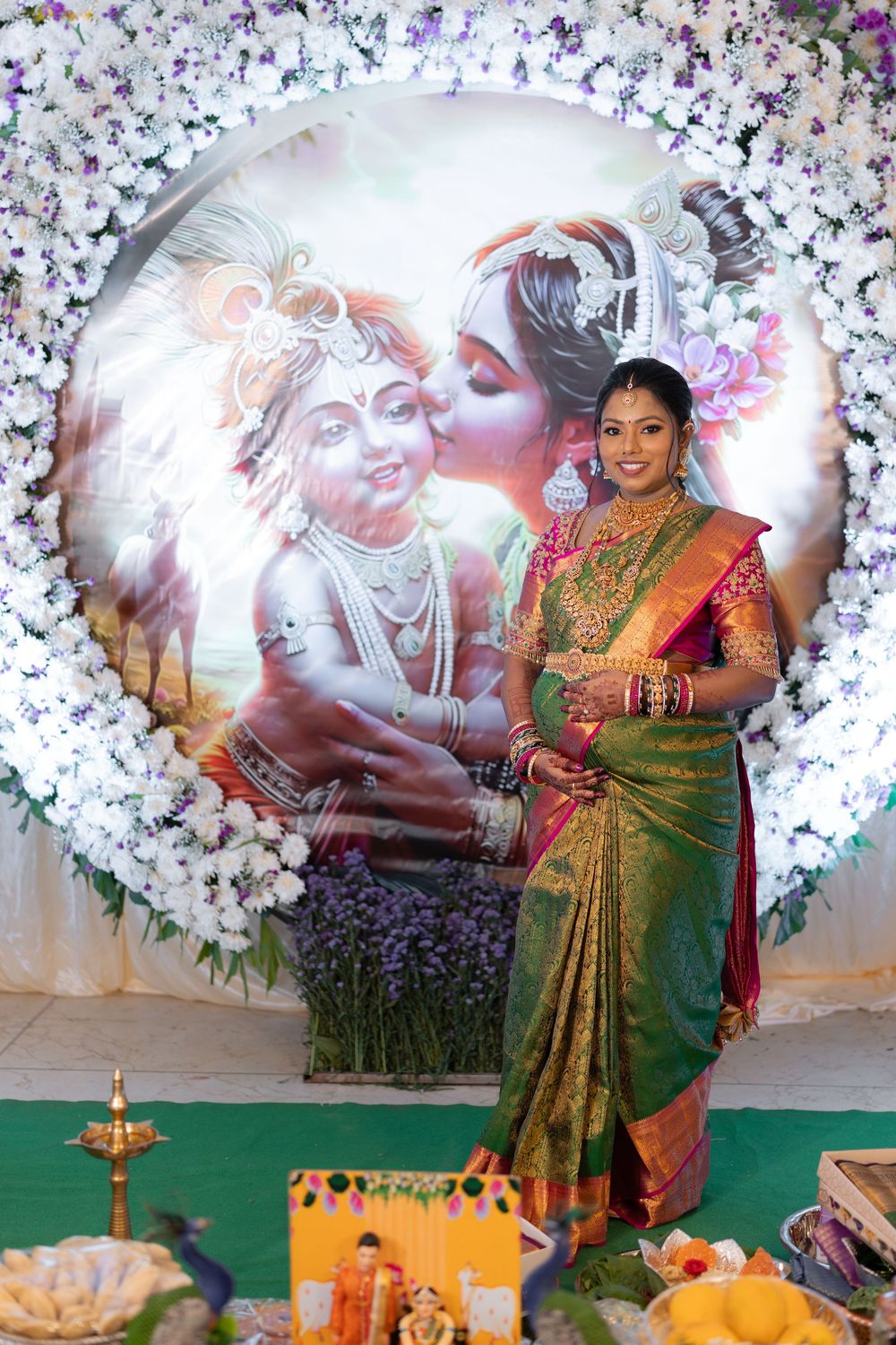 Indian bride in green Kanjivaram saree poses before a floral Krishna-Radha backdrop at a traditional Hindu wedding.