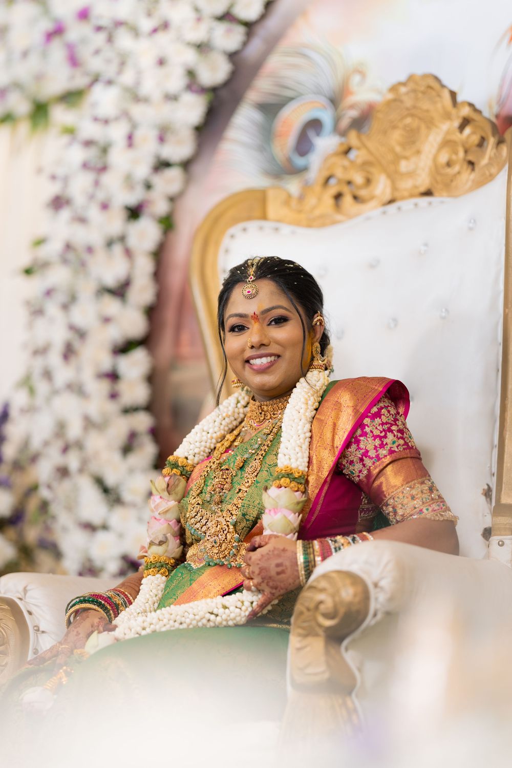 South Indian bride in silk saree and gold jewelry smiles radiantly on ornate white throne at traditional wedding ceremony.