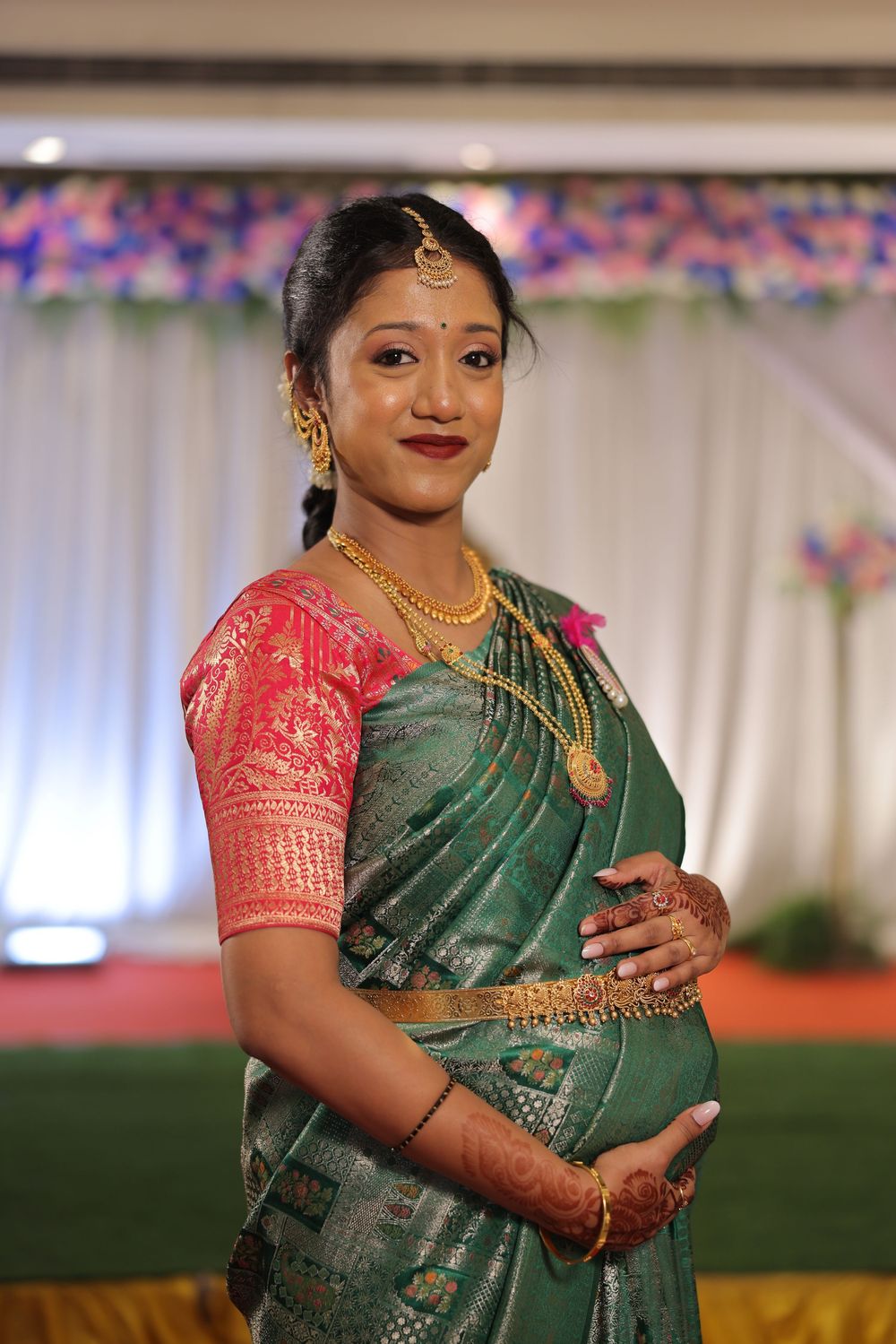 Pregnant Indian woman in green silk saree with pink blouse and gold jewelry at a traditional ceremony.