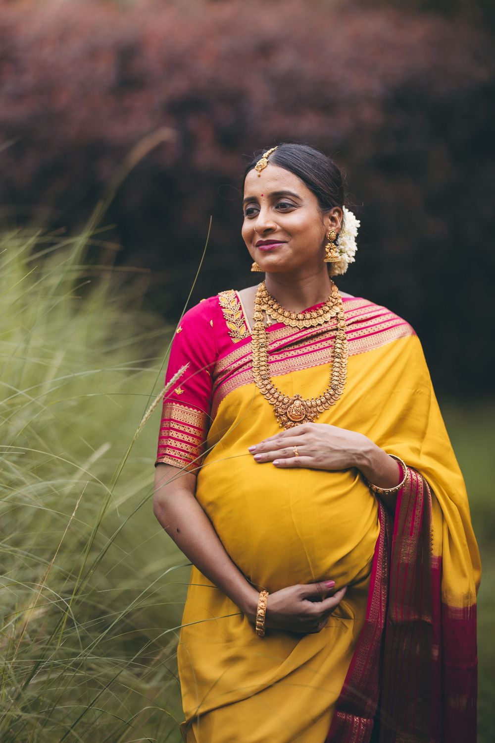 Pregnant Indian woman in yellow and red saree with gold jewelry, posing outdoors among tall grass.