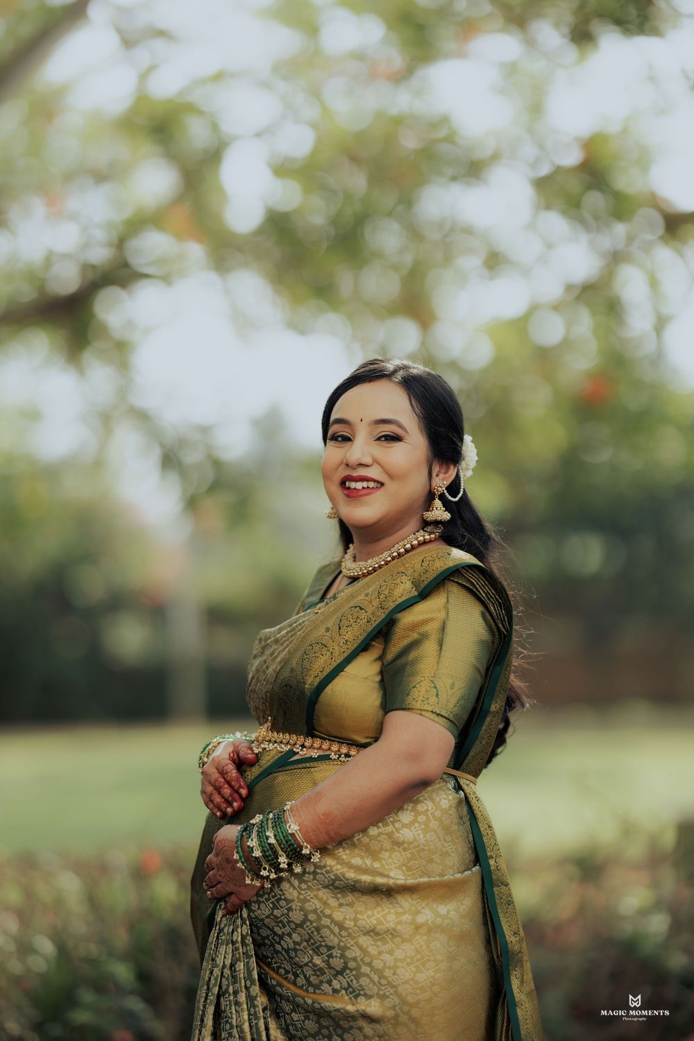 Pregnant woman in elegant green Kanjivaram silk saree with gold zari work, posing outdoors at a maternity photoshoot.