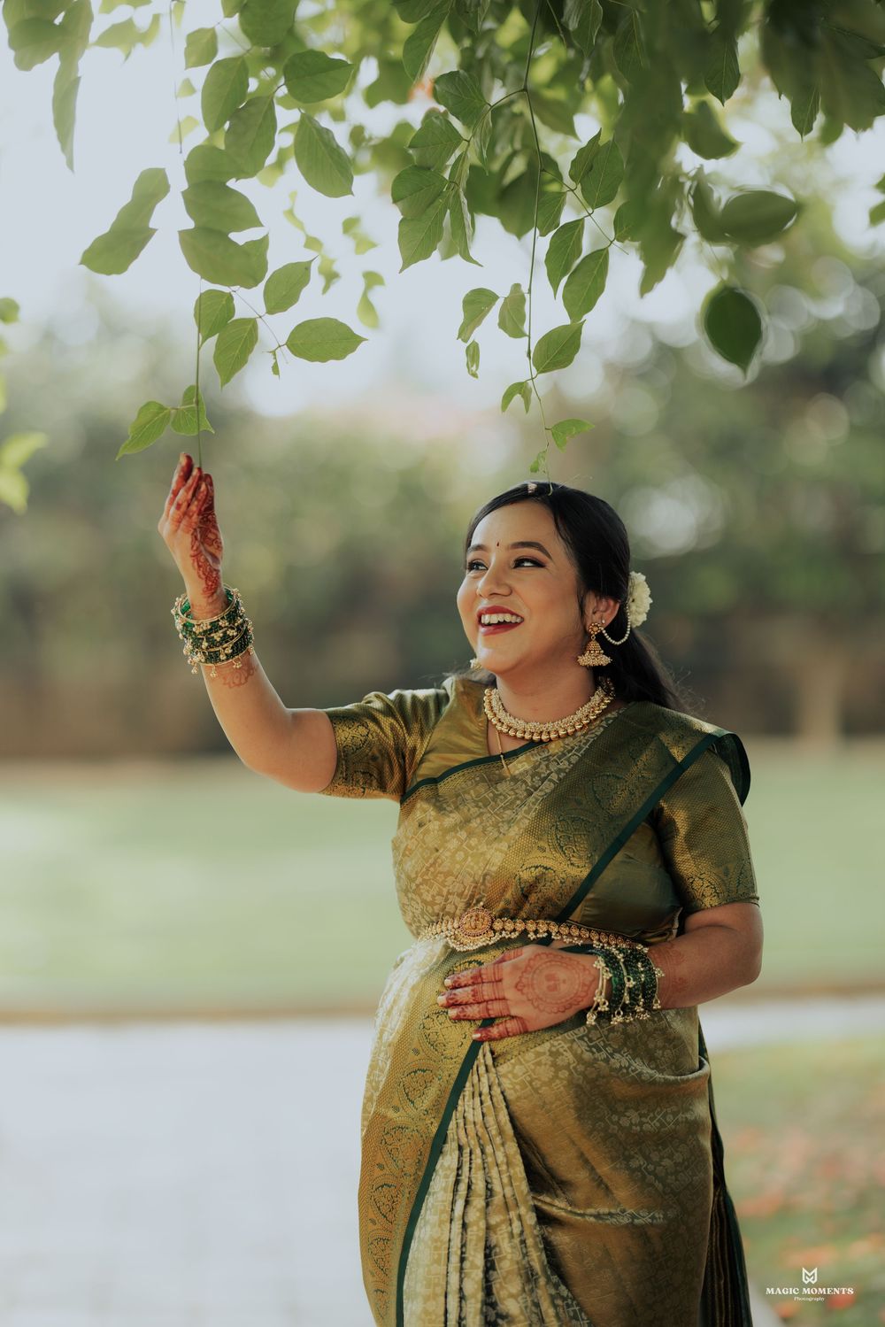 Woman in golden green saree reaching for leaves, smiling outdoors near water, adorned with traditional jewelry.