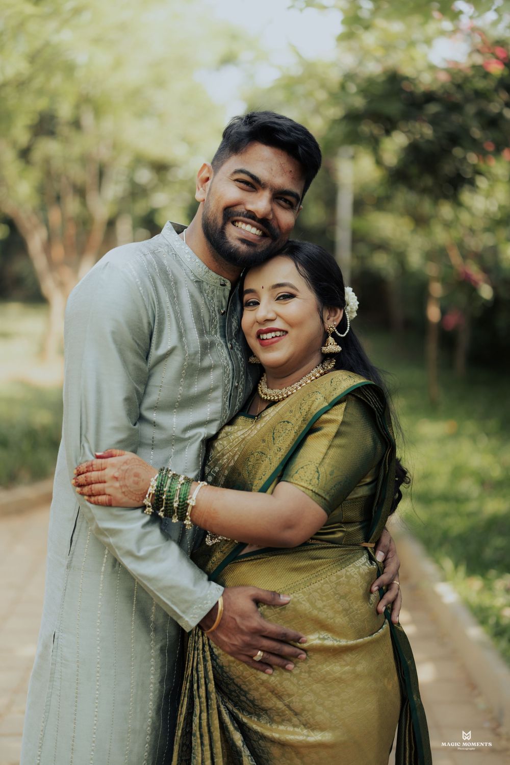 Indian couple in traditional attire embracing outdoors, man in kurta and woman in green saree with gold jewelry.