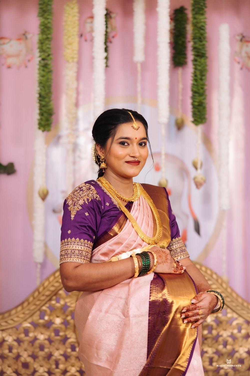 A bride in a pink silk saree with purple blouse and gold jewelry poses at a traditional South Indian wedding ceremony.