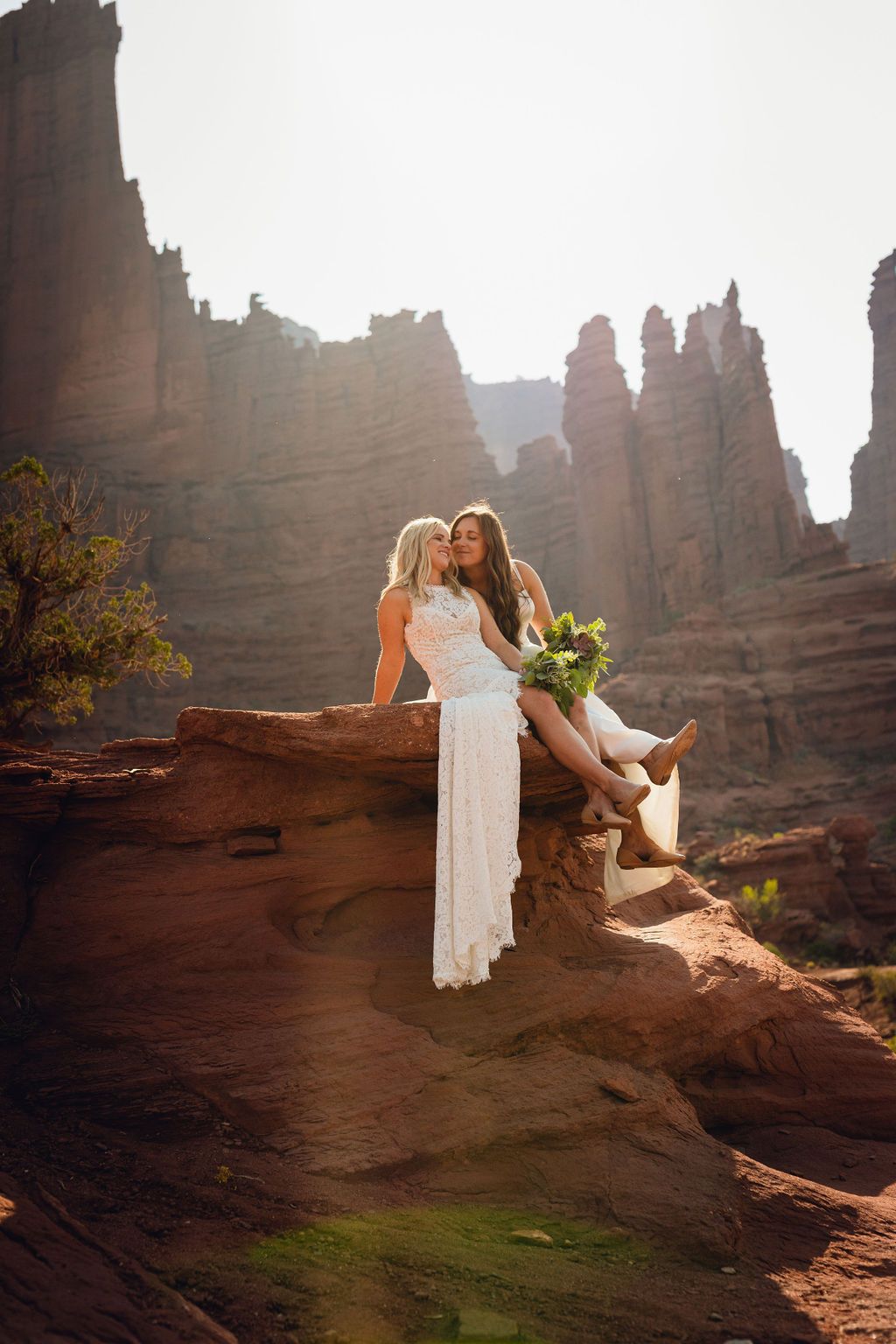 Two brides sitting on red rock formations in a desert canyon at sunset.
