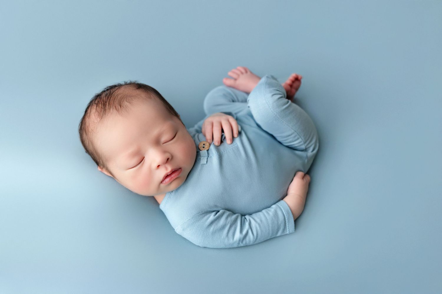 Peaceful newborn baby sleeping in a blue onesie on a soft blue backdrop in a professional photo.