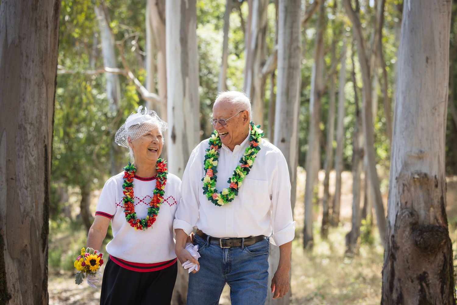 Elderly couple wearing floral leis holding hands and smiling at each other while walking through a forest.