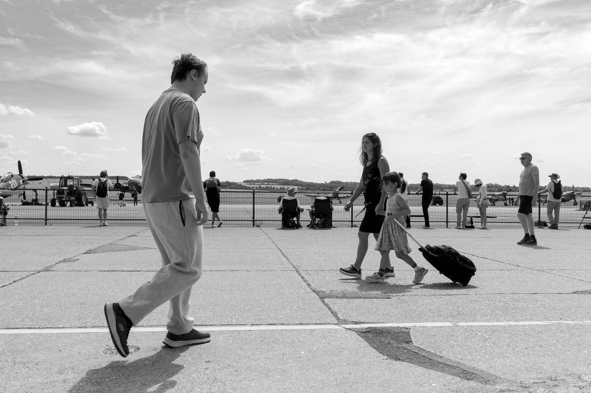 Black and white photo of people walking on an outdoor plaza, one pulling luggage, with an airfield in the background.
