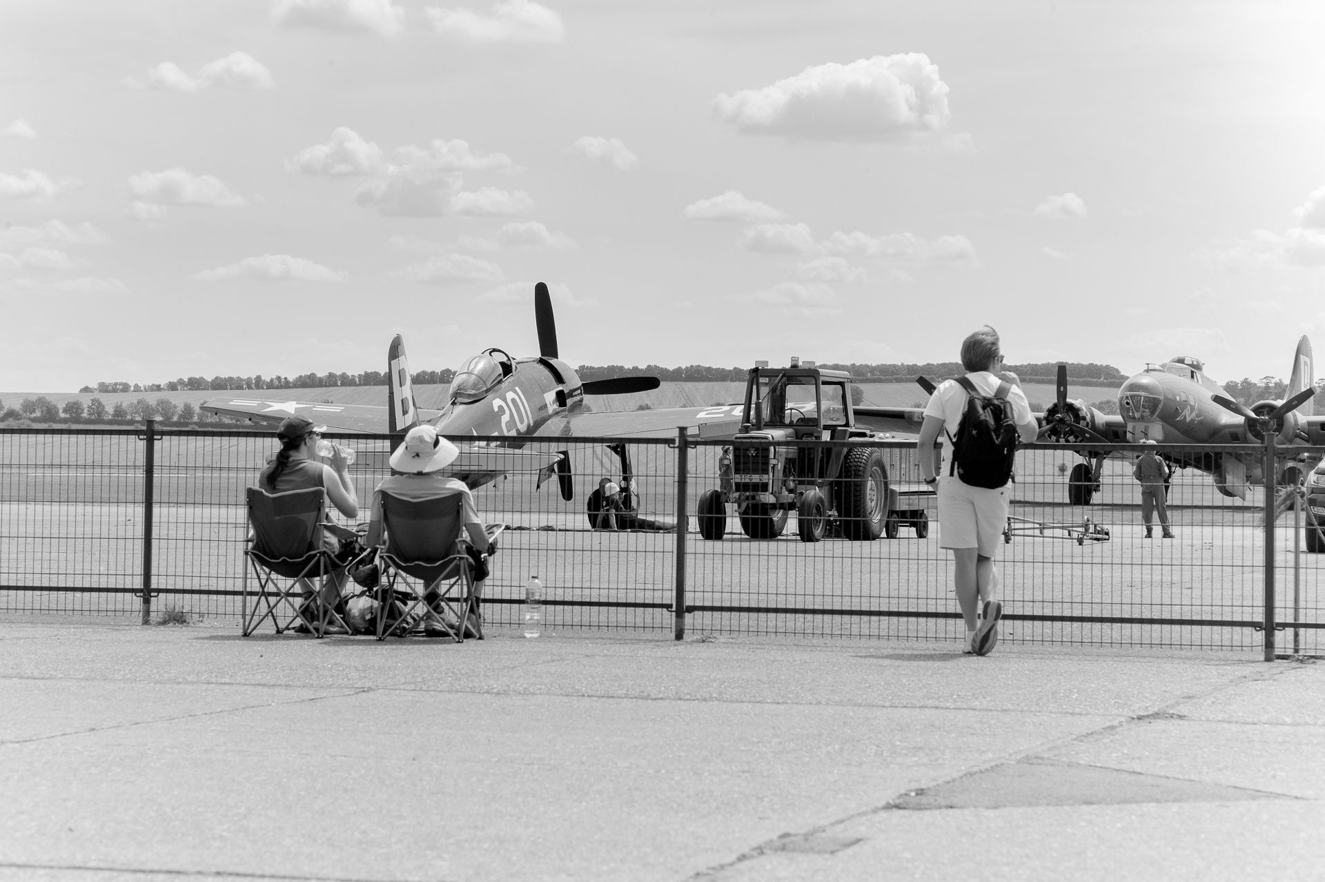 Black and white photo of people at an airshow watching vintage propeller aircraft on the tarmac behind a fence.