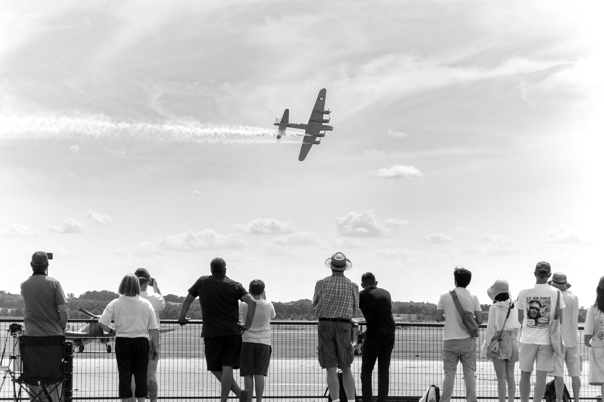 Crowd watches a vintage WWII bomber aircraft fly overhead at an outdoor airshow in black and white photography.