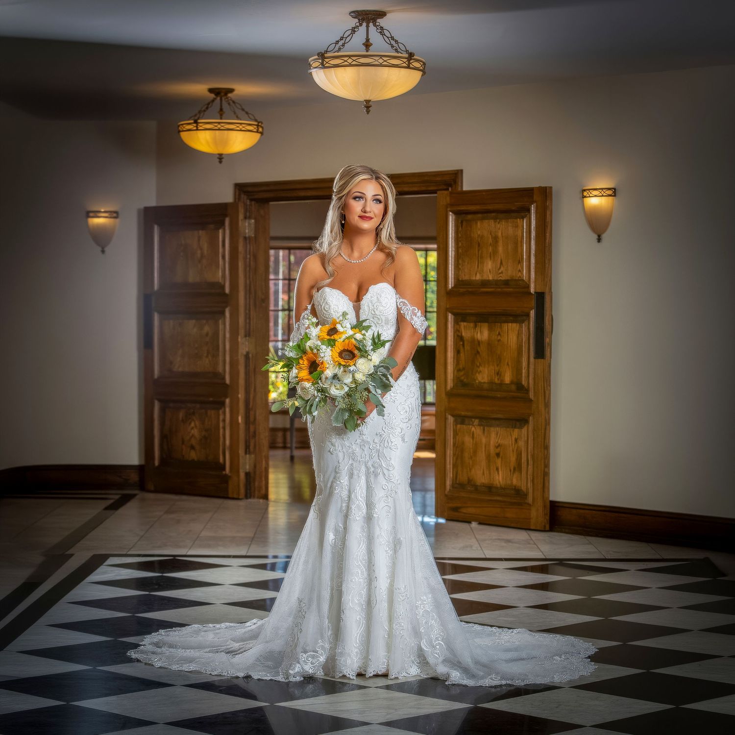 Bride in white lace mermaid gown holding sunflower bouquet stands before wooden doors in elegant venue.