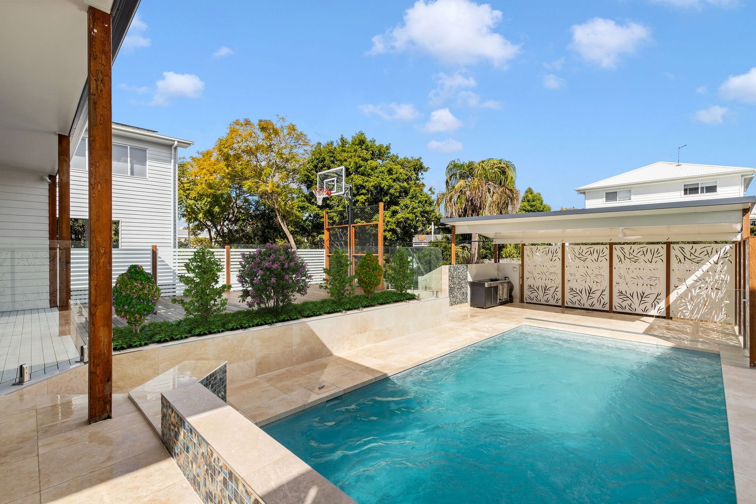 Bright outdoor pool area with timber pergola, decorative screen fence, and basketball hoop.