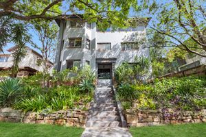 A charming multi-story white apartment building surrounded by lush tropical gardens and stone retaining walls on a sunny day.