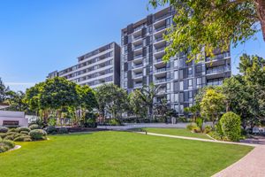 Modern multi-story apartment complex with lush green lawn and manicured gardens under a clear blue sky.