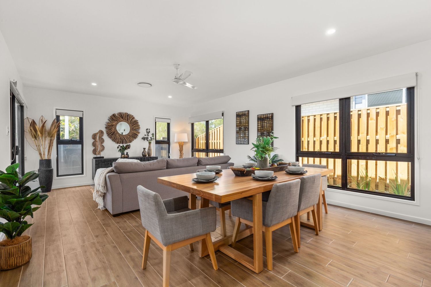 Open-plan dining room with timber table, grey chairs, ceiling fan, and large windows overlooking garden.