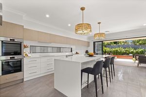 Contemporary white kitchen with large island bench, gold chandeliers, double ovens, and outdoor views.