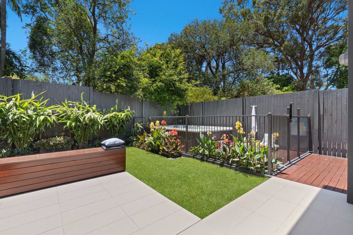 Landscaped backyard with artificial grass, colorful garden beds, timber deck, and pool area.