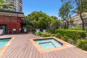 Outdoor spa and timber deck area beside a pool, surrounded by tropical gardens and apartment buildings.