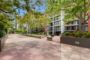 Tree-lined pedestrian courtyard with garden beds and low-rise apartment buildings in an urban complex.