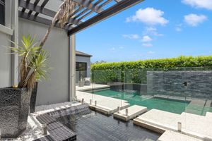 Modern outdoor pool area with glass fencing, pergola, and manicured hedge under blue sky.