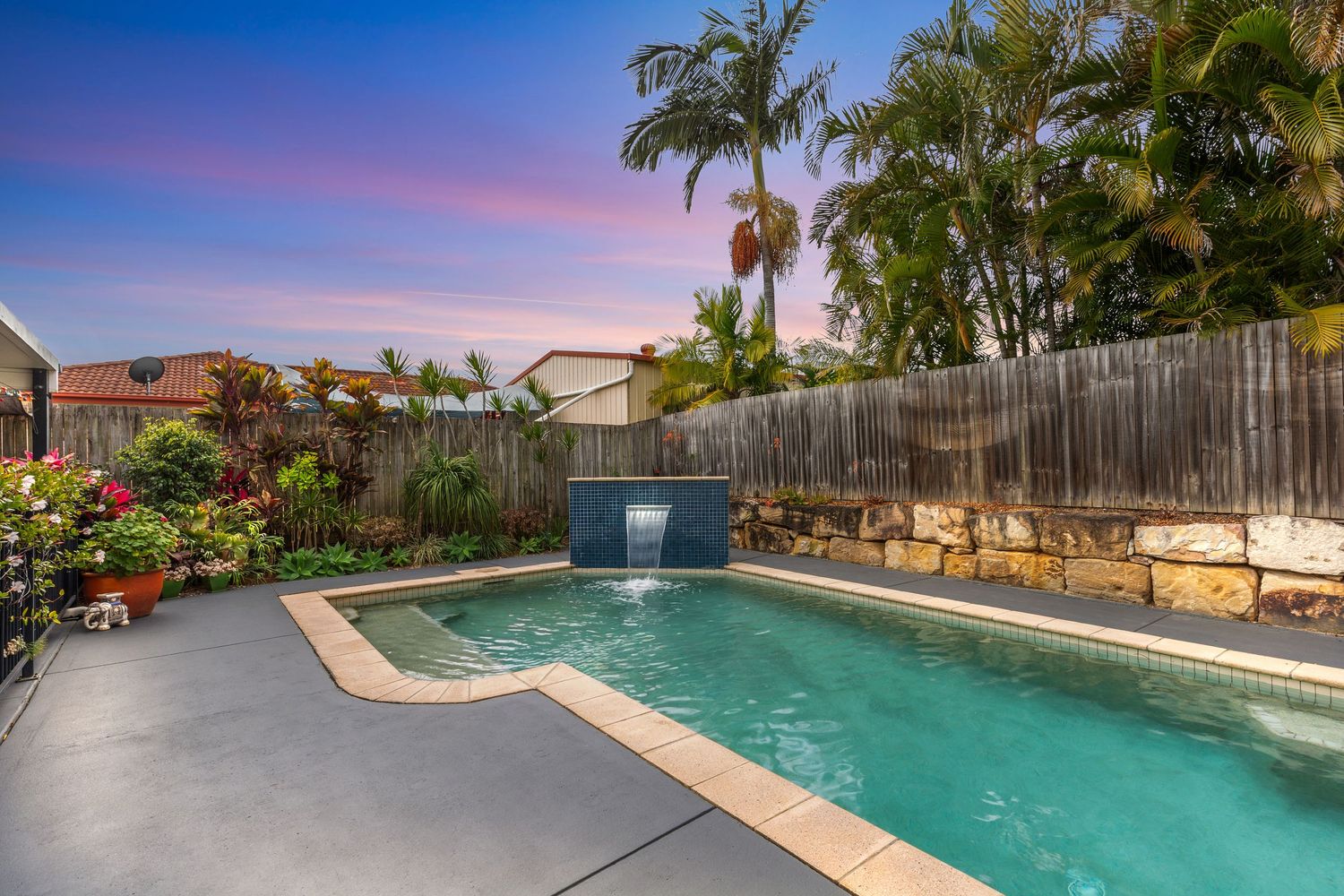Backyard pool with waterfall feature, tropical plants, and wooden fence at sunset.