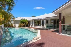 Luxury home with sparkling pool, timber deck, glass balustrade, and covered outdoor entertaining area under blue skies.