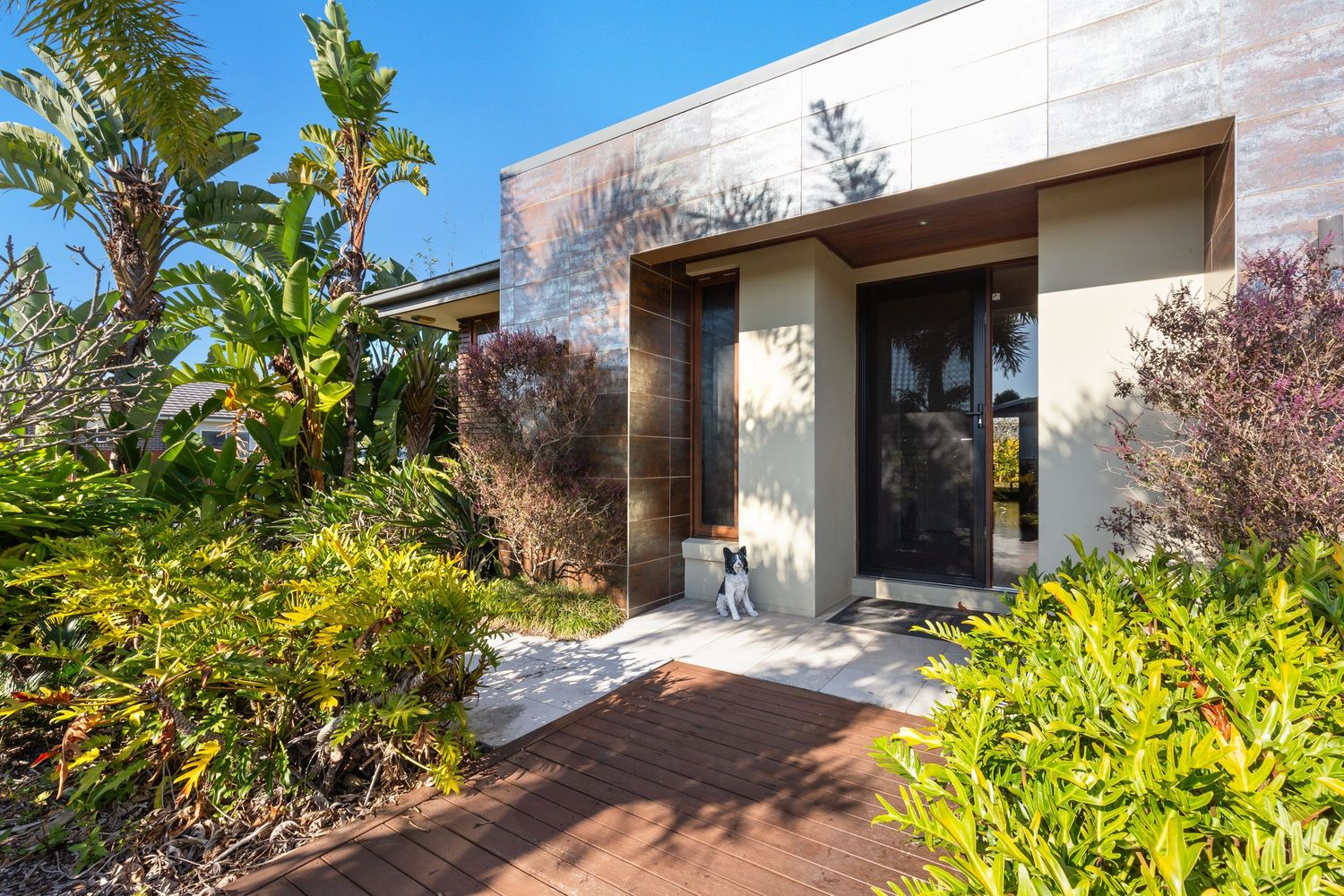 Contemporary home entrance with timber pathway, lush tropical gardens, and modern stone facade.