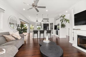 Elegant white living room with dark hardwood floors, fireplace, ceiling fan, and open kitchen view.