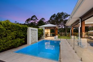 Luxury backyard pool with glass fencing, water feature wall, and outdoor gazebo at dusk.