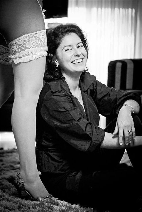 Black and white photo of a smiling woman sitting on the floor next to a leg wearing a lace garter stocking.
