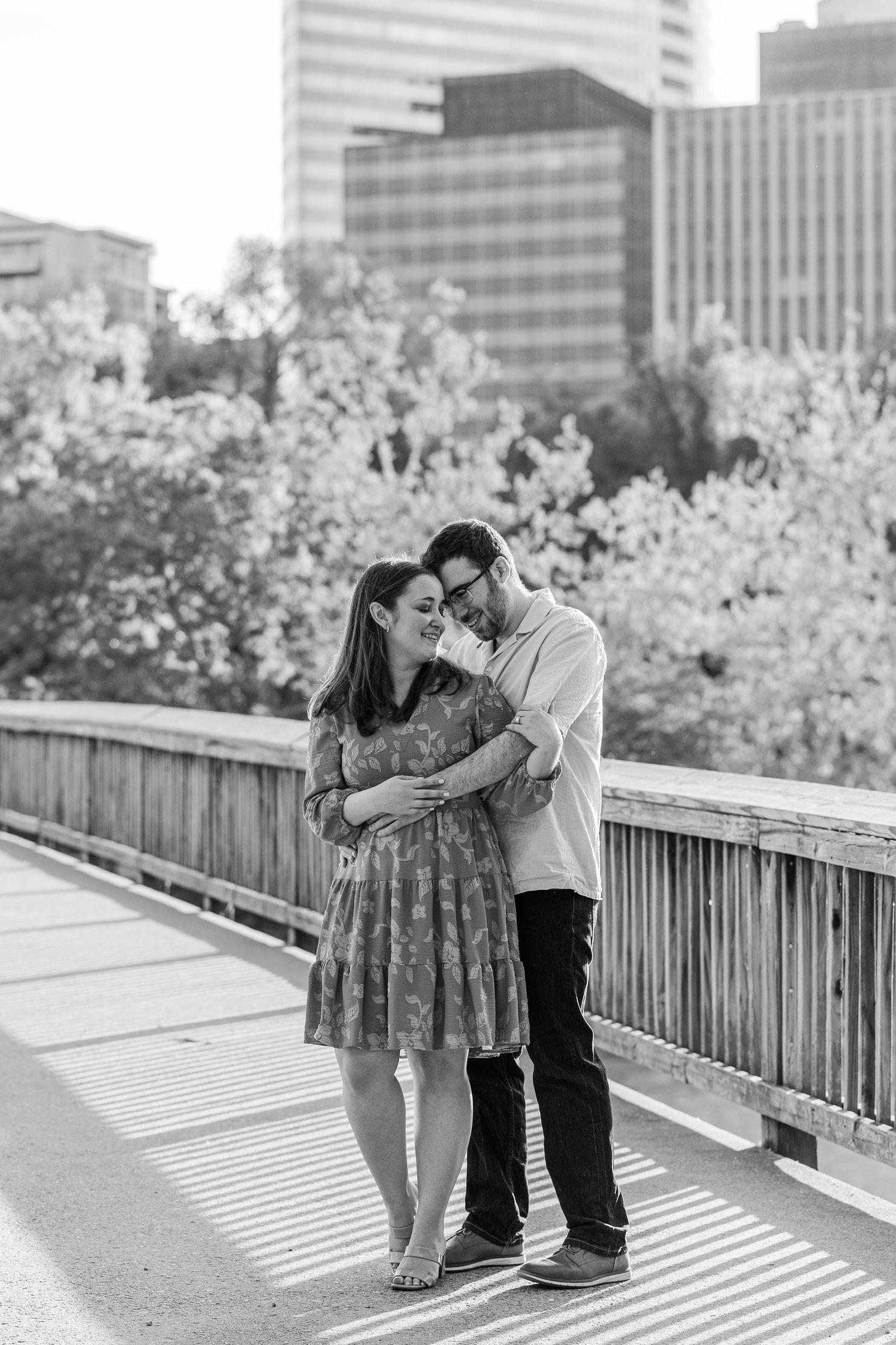 Black and white photo of a couple embracing on a bridge with city skyline and blooming trees in the background.