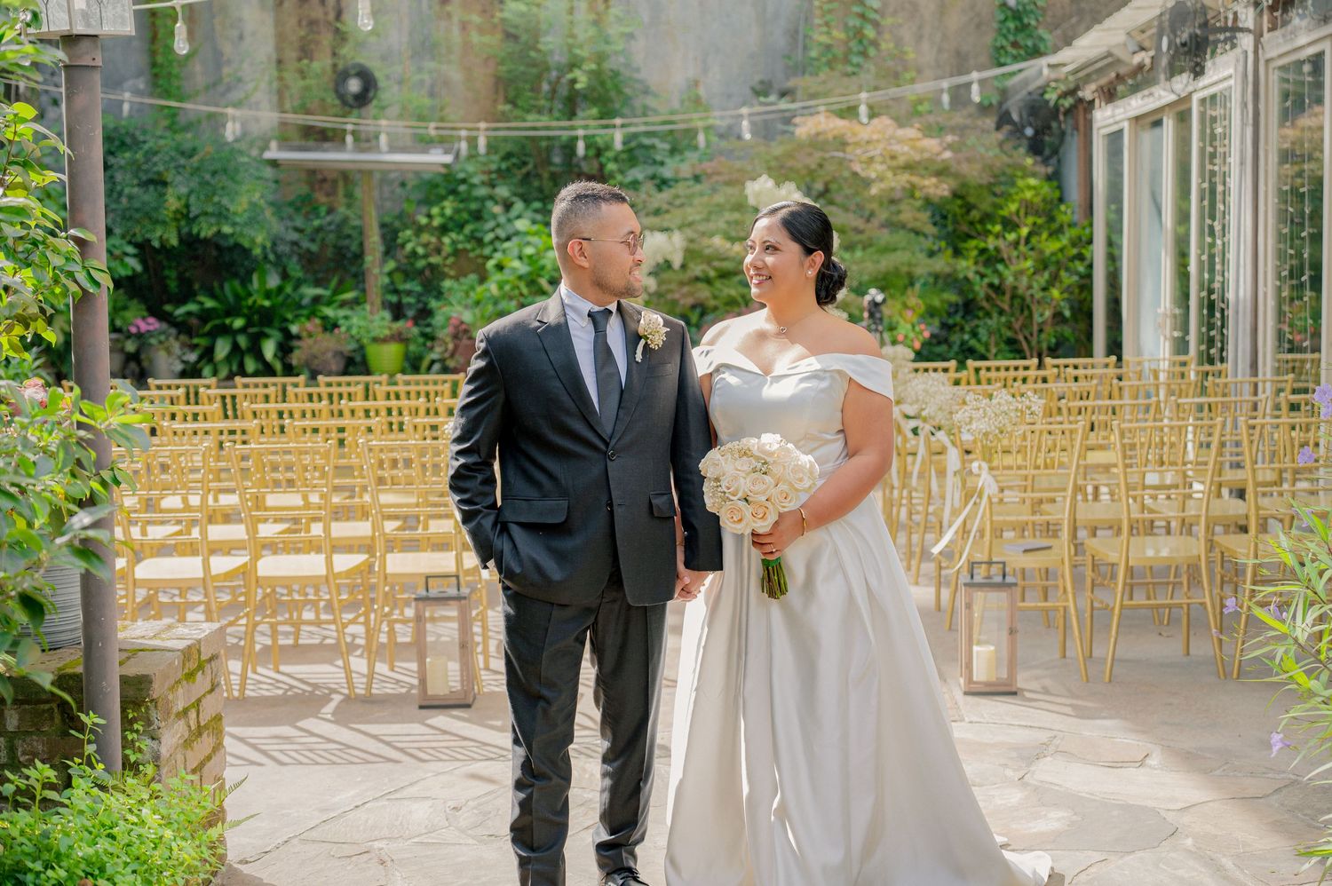 Bride and groom stand together in an elegant outdoor wedding venue with gold chairs and lush greenery backdrop.