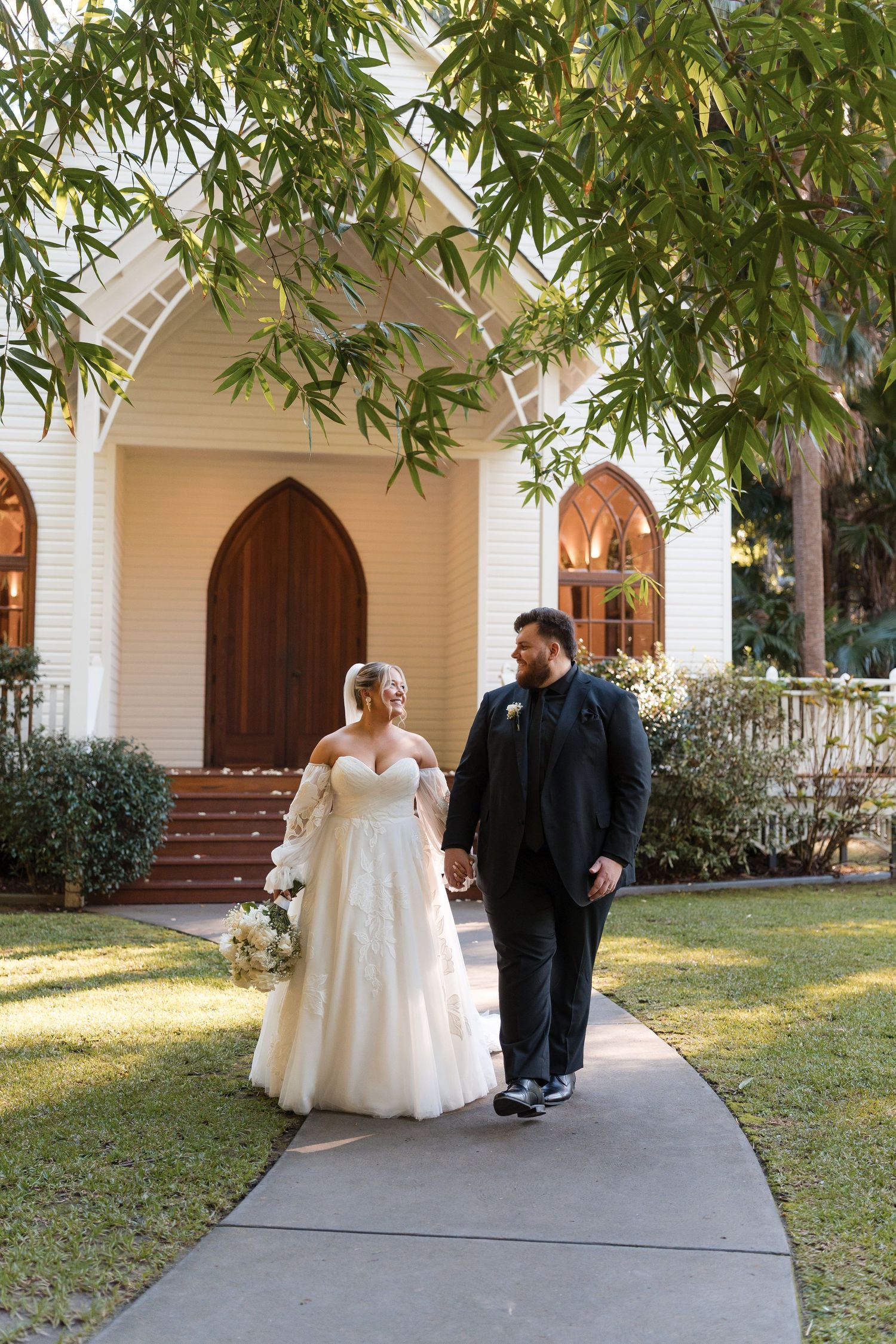 Bride and groom walk hand-in-hand on a church pathway, surrounded by lush greenery on their wedding day.