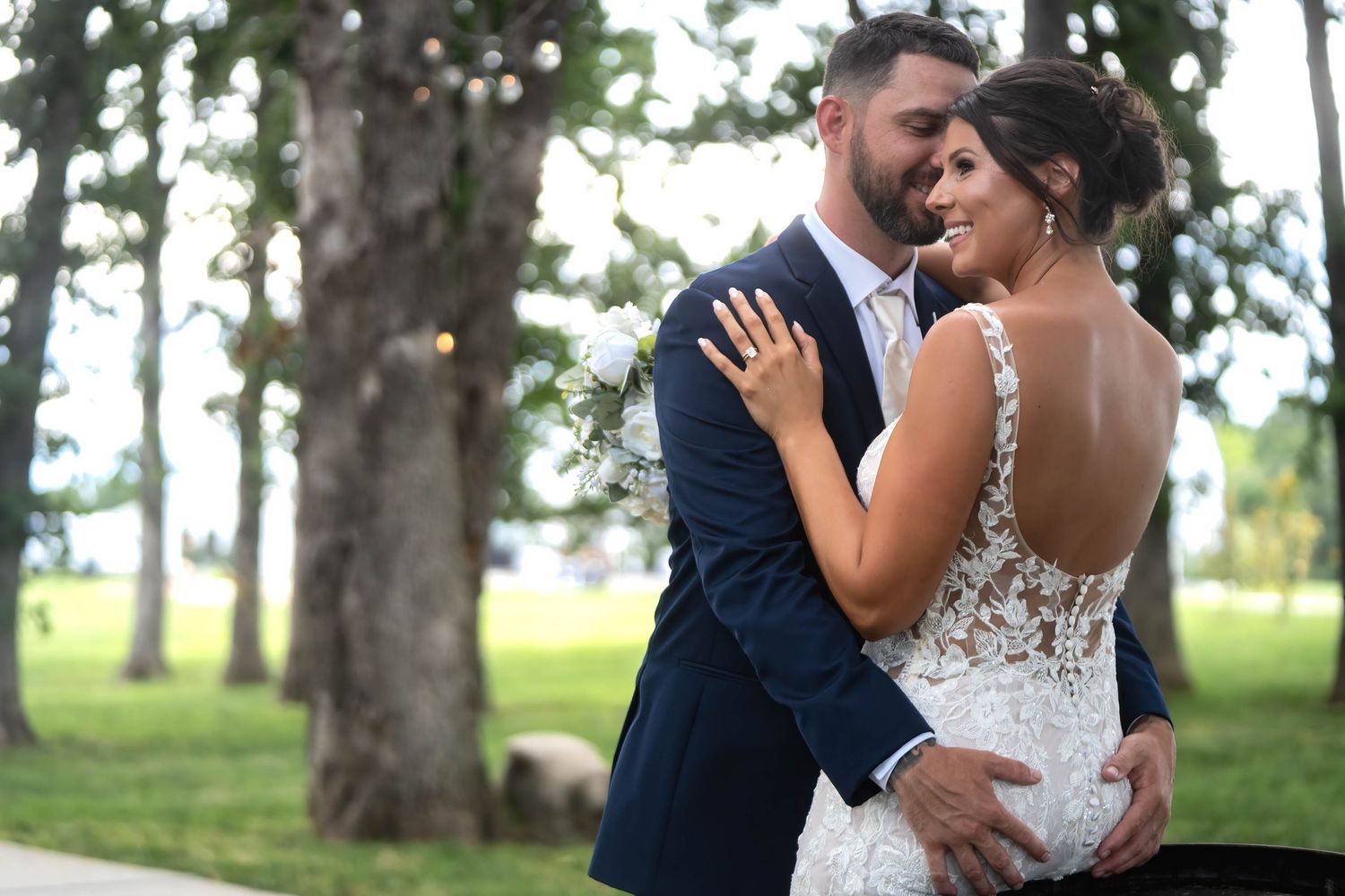A bride and groom embrace outdoors among tall trees, the bride wearing a lace backless gown and the groom in a navy suit.