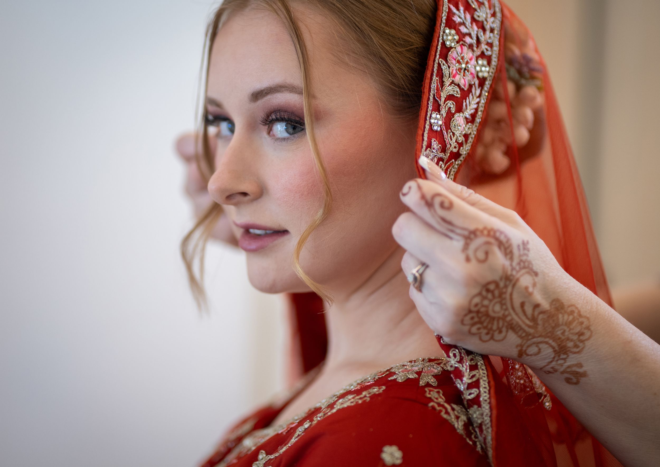 A bride in red Indian bridal attire with mehndi henna hands adjusting her embroidered dupatta veil.