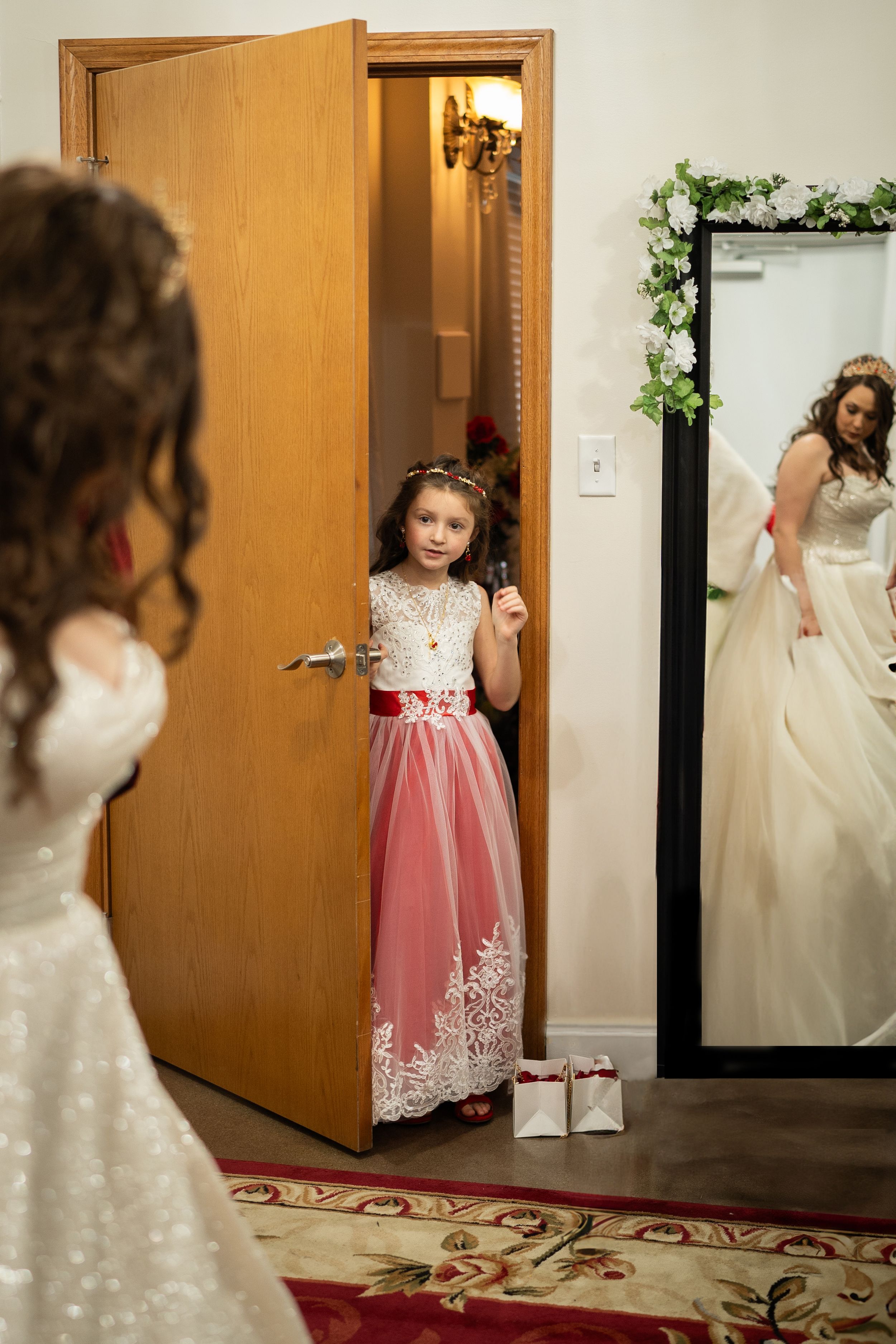 Young flower girl in pink and white lace dress stands in doorway as bride prepares in mirror-adorned dressing room.
