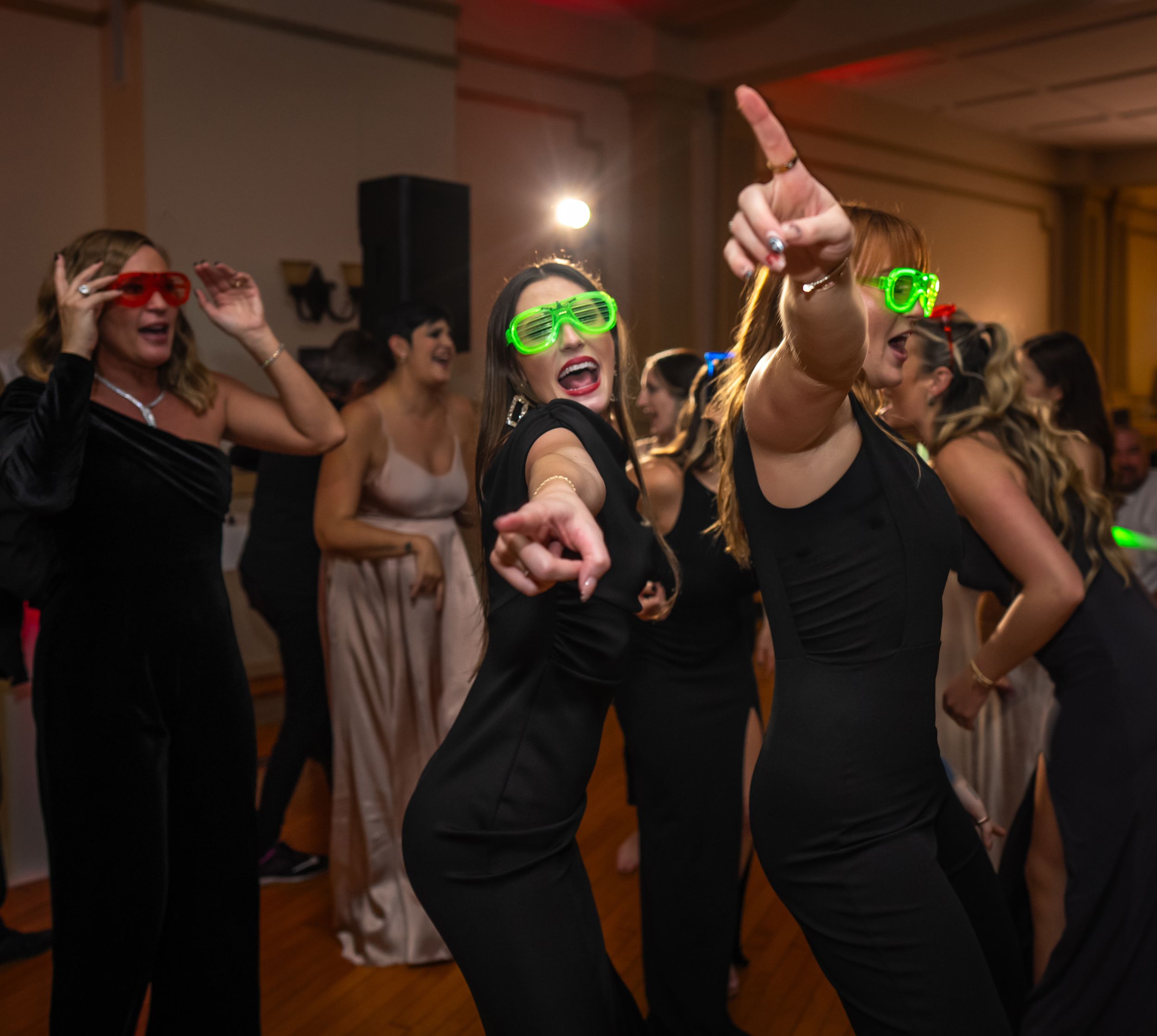Women in black outfits dancing at a party, wearing neon green novelty glasses and glow accessories.