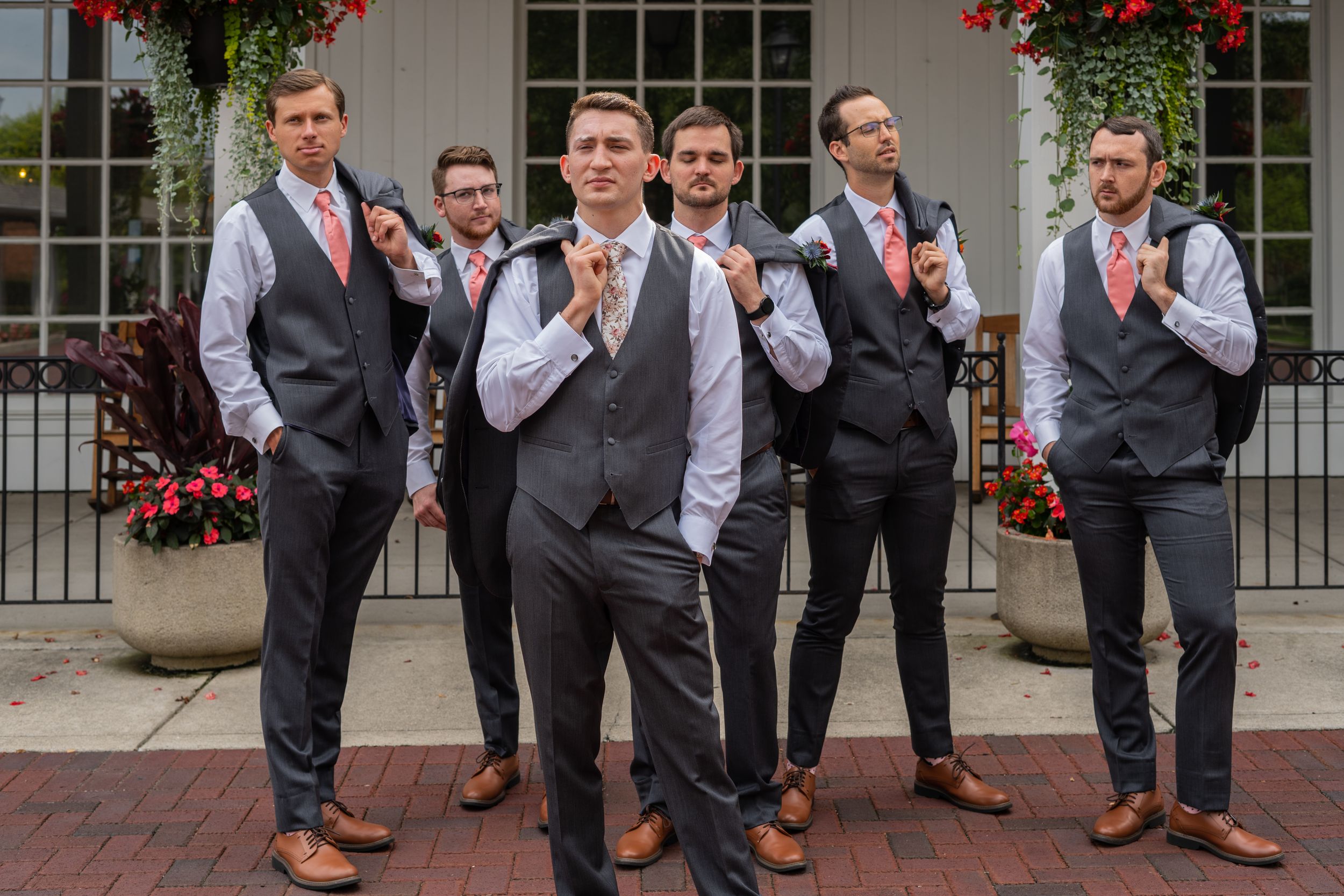 Groom and groomsmen in gray vests and coral ties pose playfully outside a white building with red flowers.