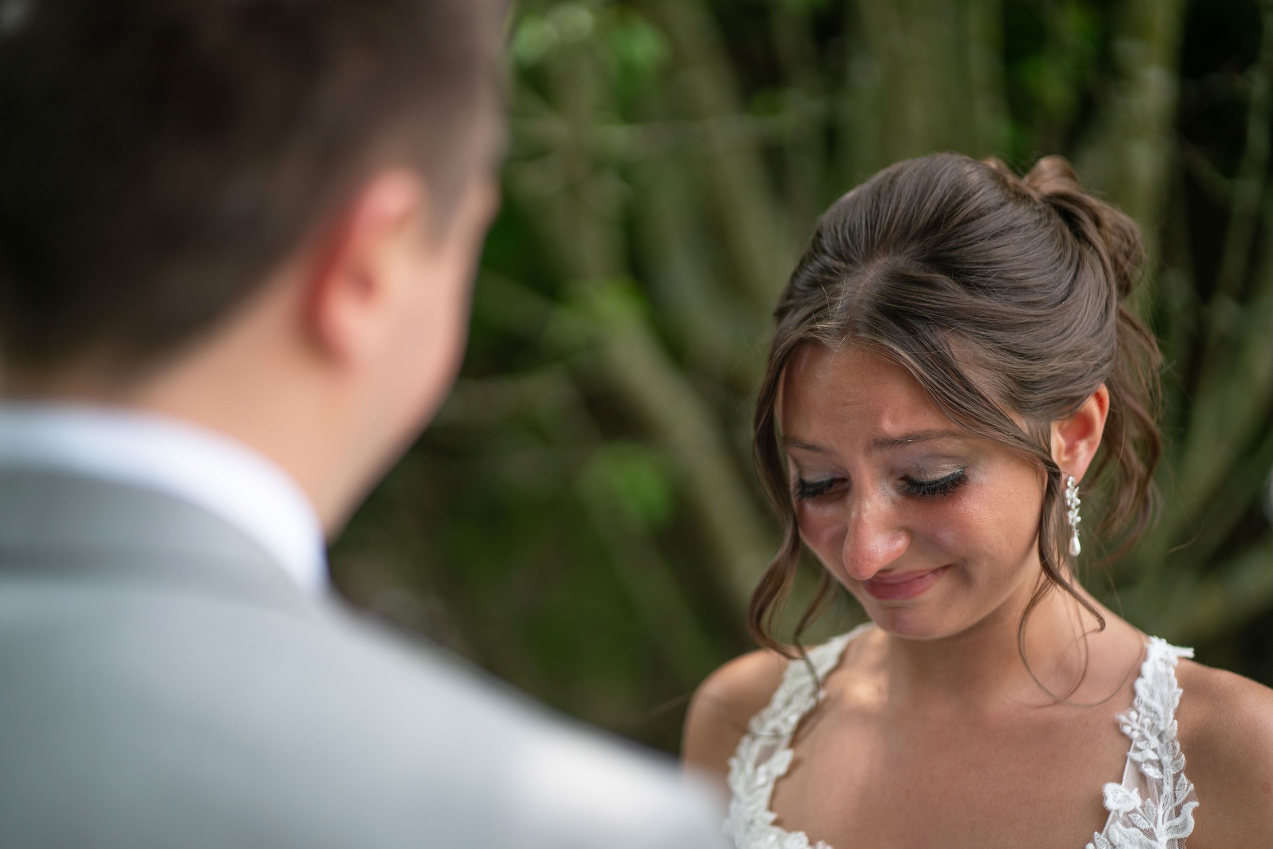 Emotional bride crying tears of joy during outdoor wedding ceremony, wearing lace dress and elegant updo hairstyle.