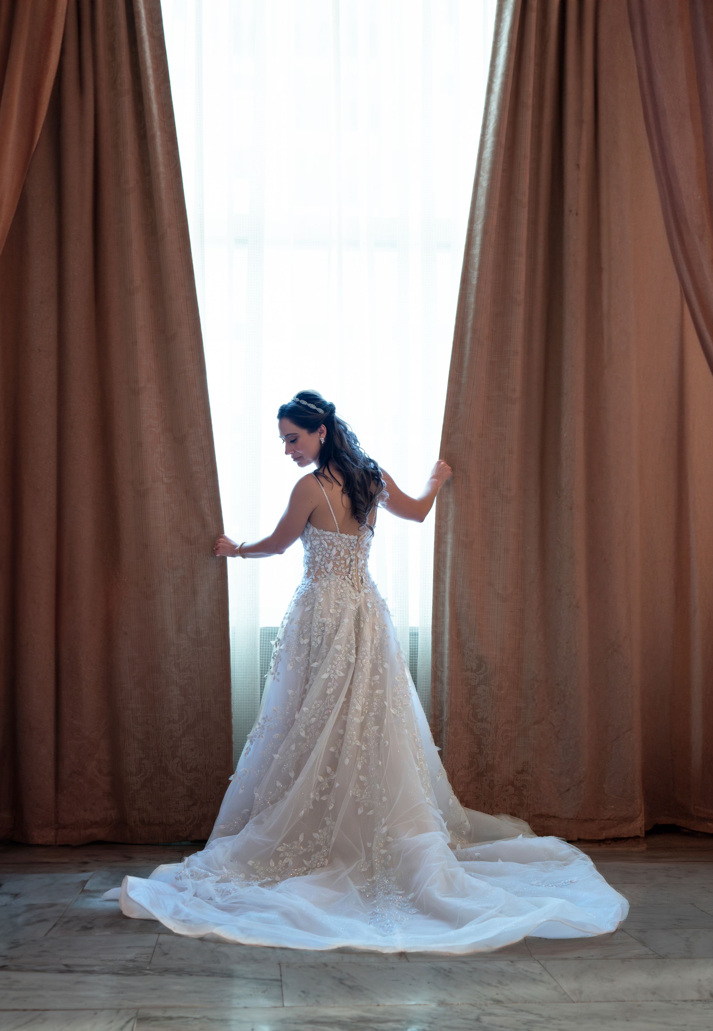 Bride in a backless floral lace wedding gown with a train stands by a sunlit window framed by elegant drapes.