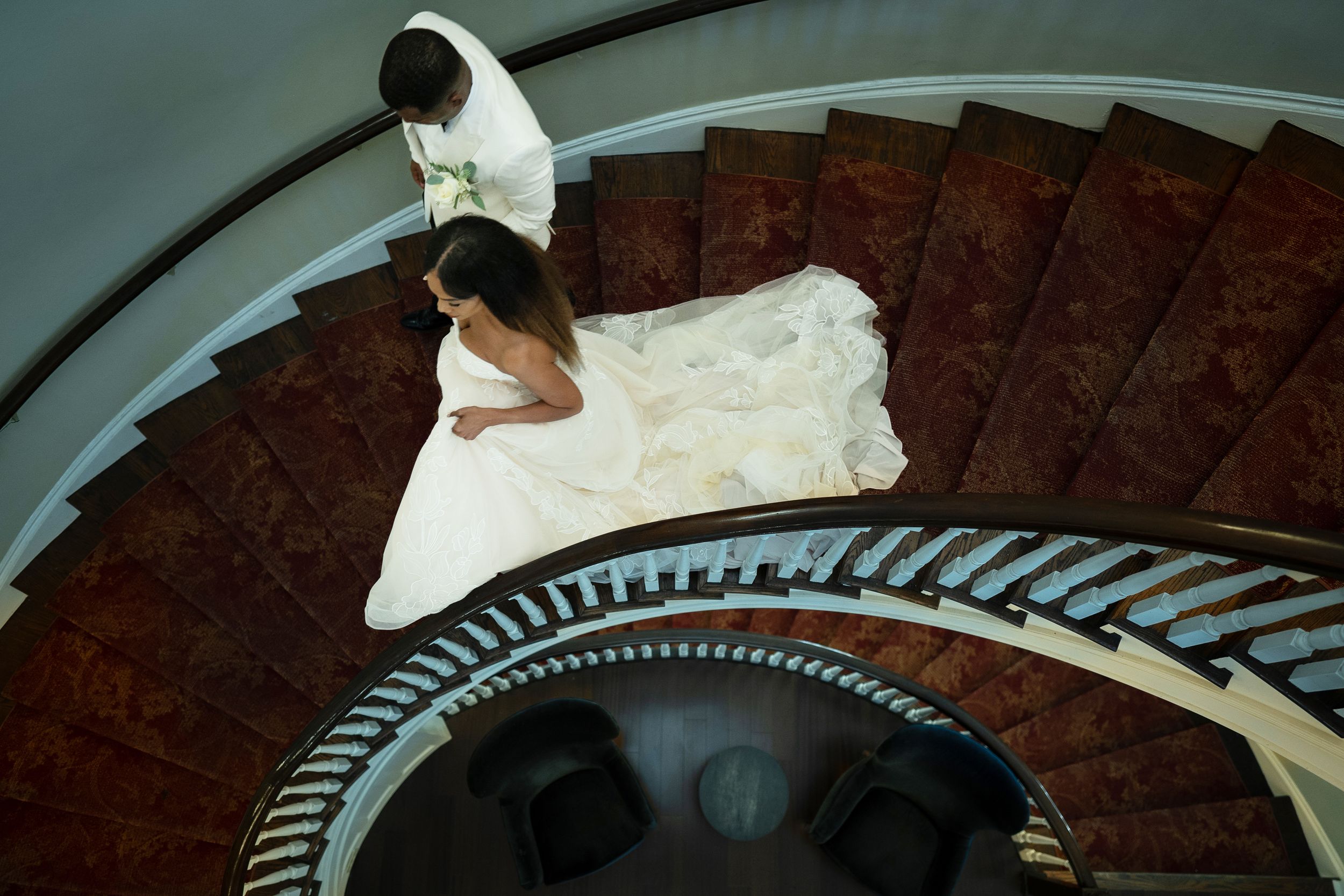 Aerial view of bride and groom on elegant spiral staircase, bride in flowing white gown, captured from above.