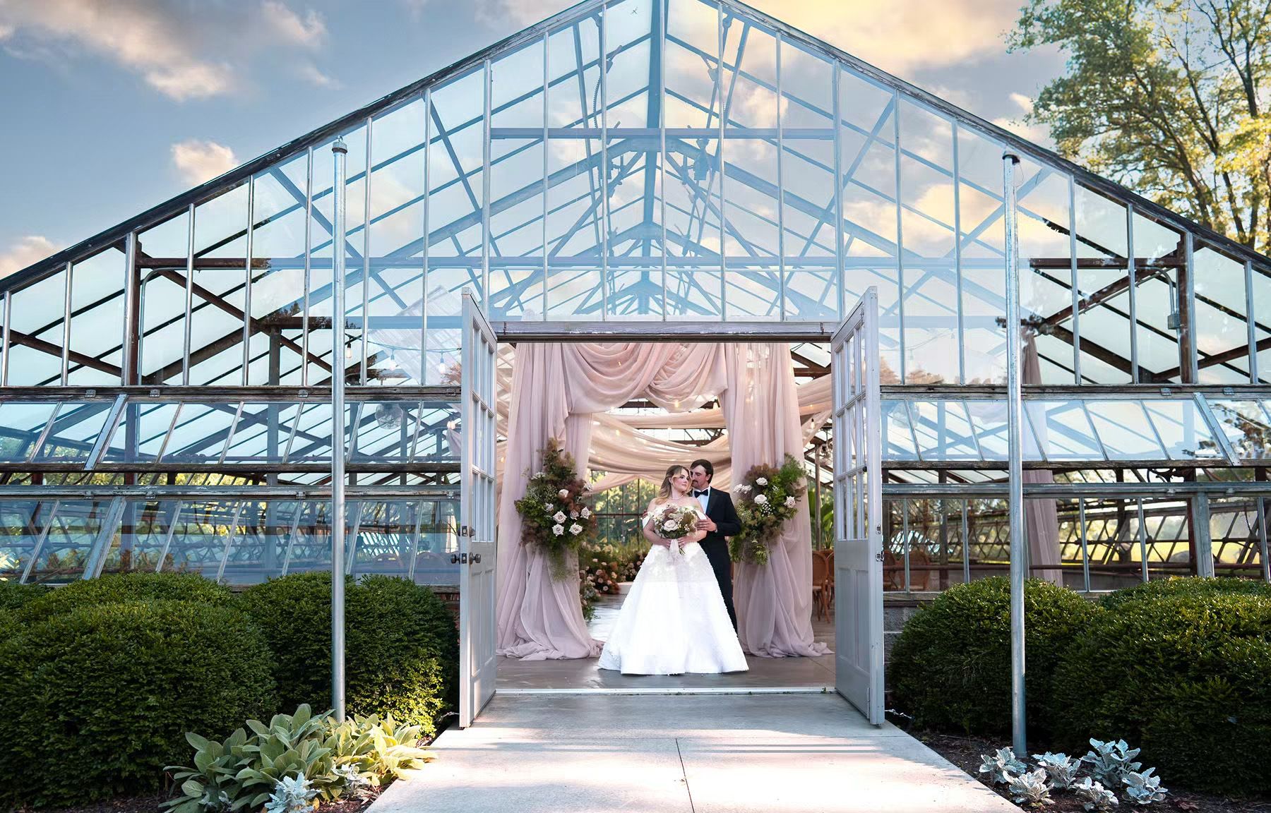 Bride and groom kissing under floral arch in elegant glass greenhouse wedding venue at dusk.