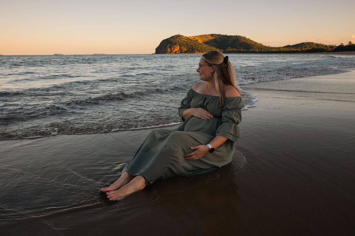 Pregnant woman in sage green off-shoulder dress sitting on sandy beach at sunset with ocean waves and green headland.