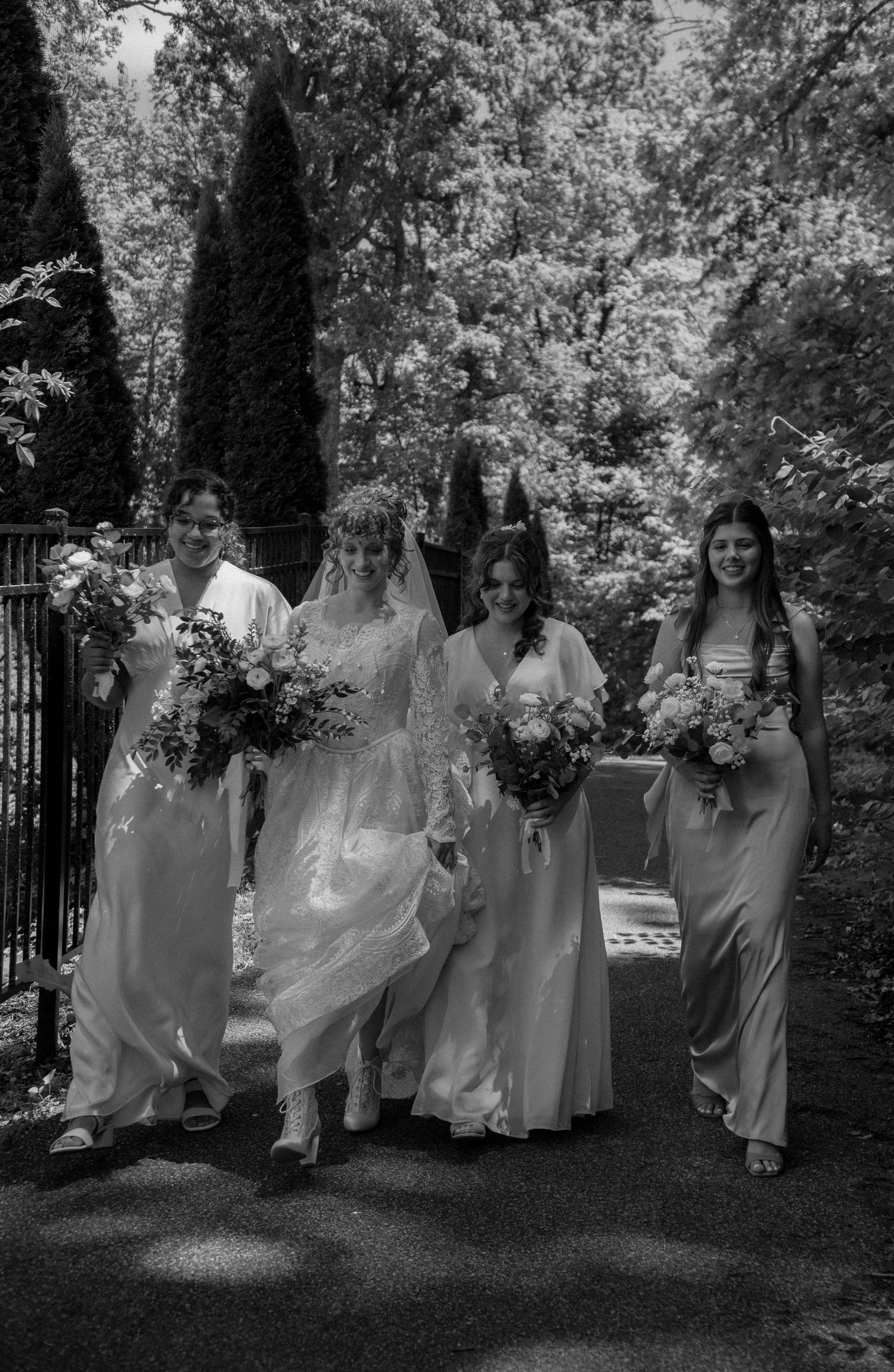 Bride in white gown walks with three bridesmaids in pastel dresses, holding bouquets on a tree-lined path.