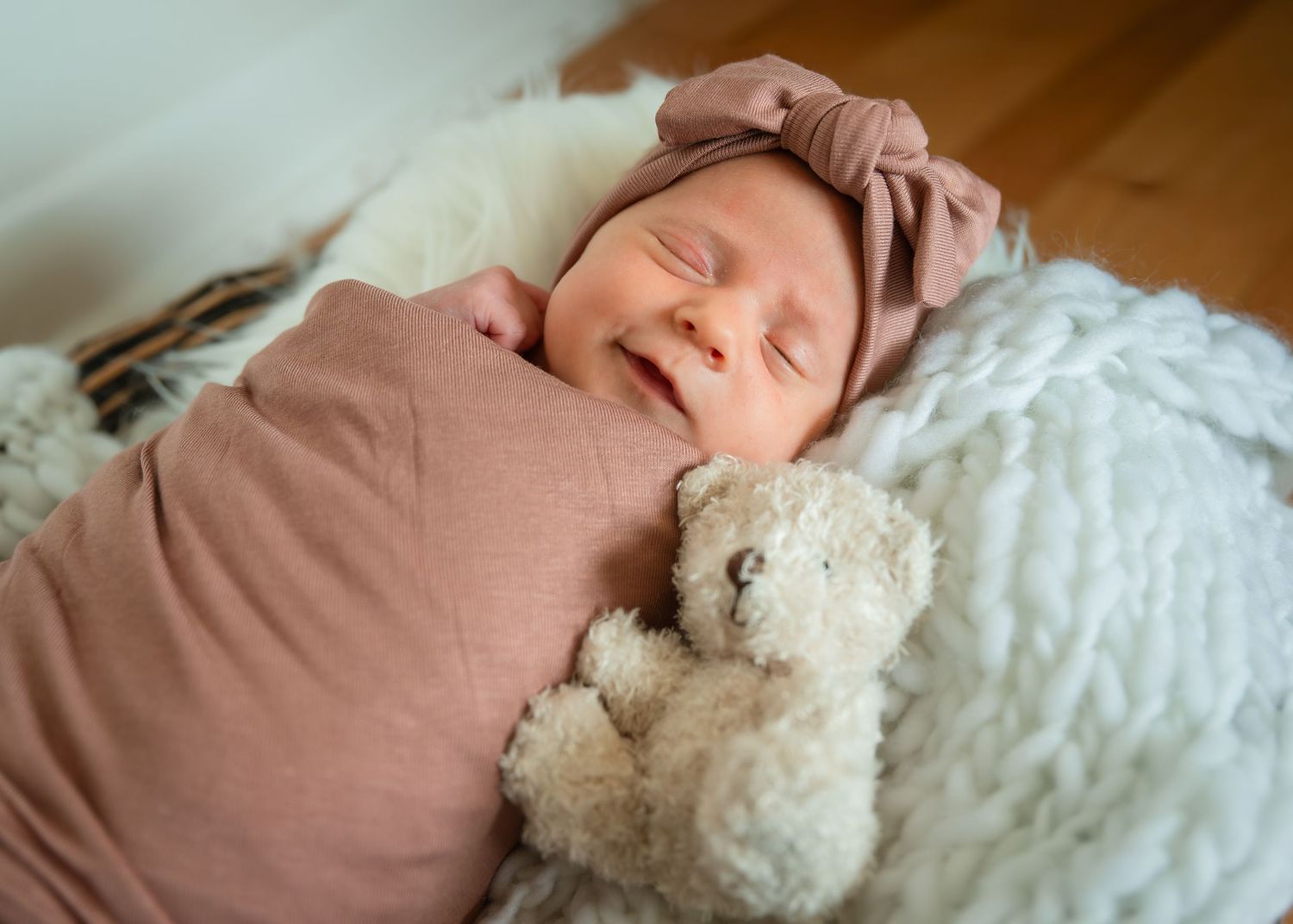 Smiling newborn baby wrapped in pink swaddle with teddy bear, wearing bow headband, sleeping on white fluffy blanket.