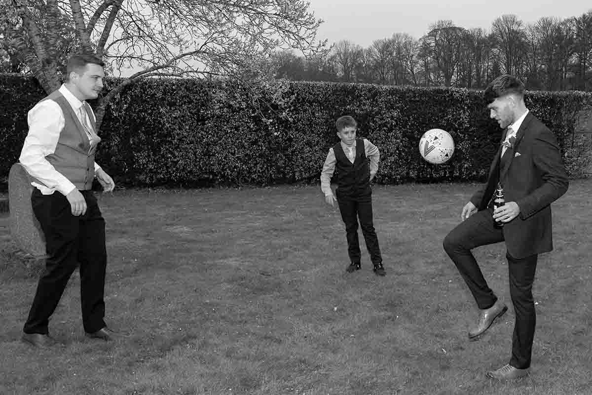 Three men in formal suits playing football on a garden lawn in a black and white wedding photo.