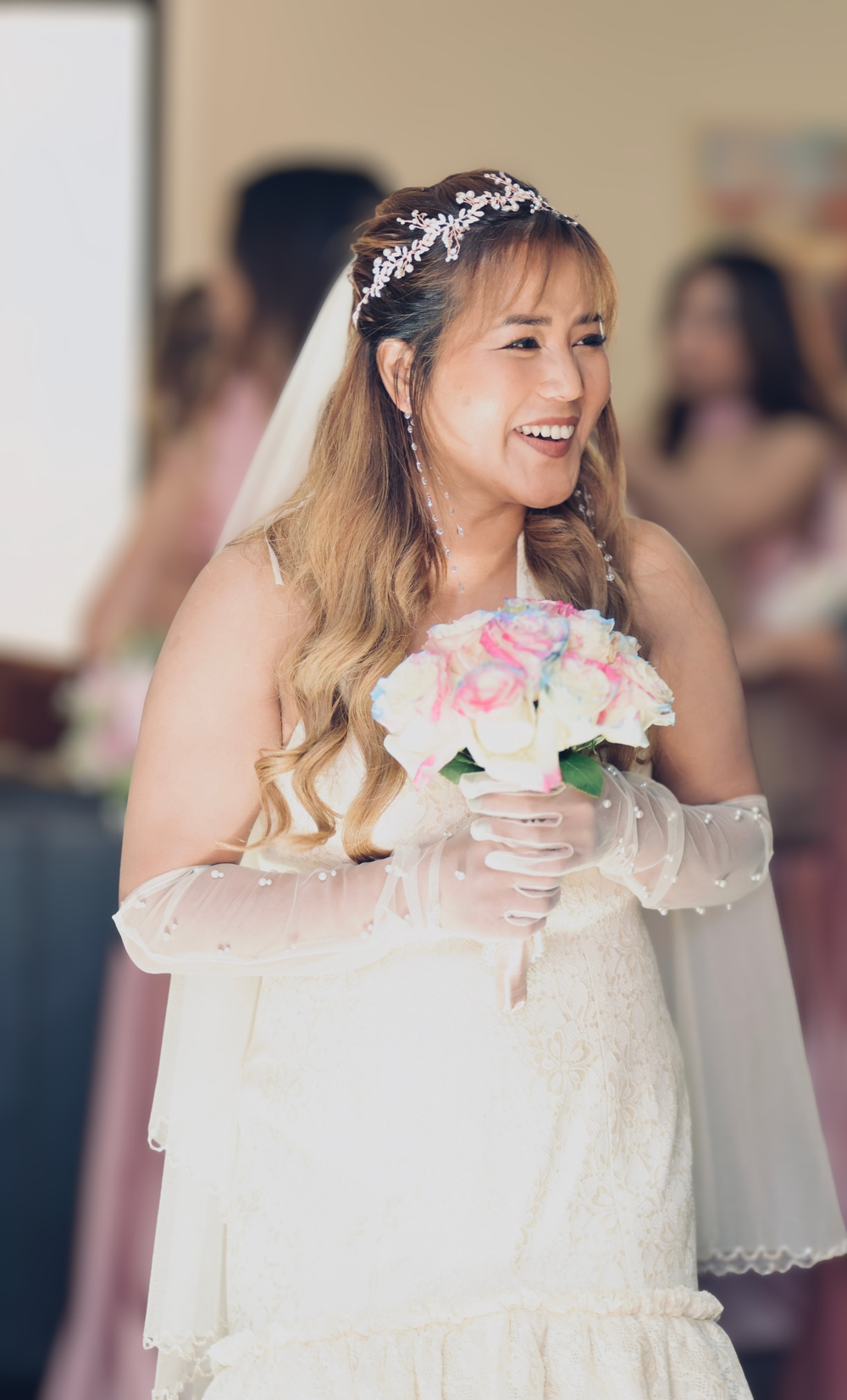 Smiling bride in white off-shoulder gown holding pink and white bouquet, wearing veil and floral headpiece.