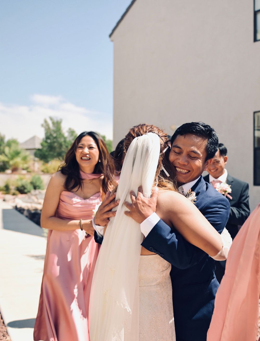 Groom in navy suit embraces bride in white wedding dress and veil outdoors while bridesmaid in pink looks on.